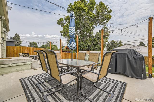 Patio dining set on striped rugs beside a covered grill and wooden fence.