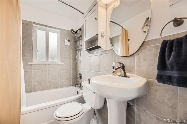 Bathroom with gray tile, a bathtub and shower, and a pedestal sink beneath a round mirror.