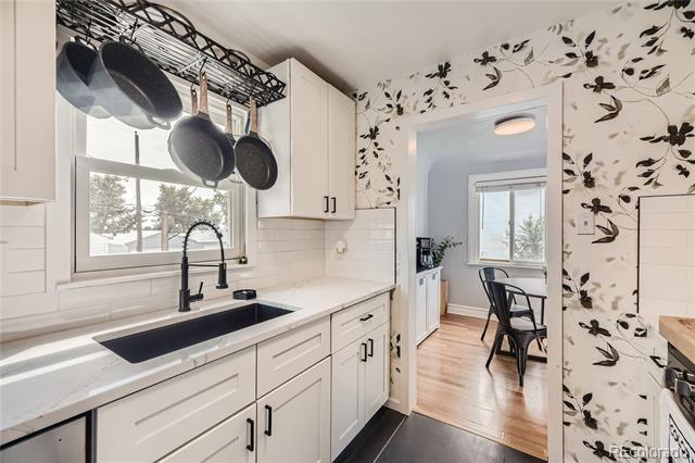 Kitchen with white cabinets, black farmhouse sink, hanging pots, and floral wallpaper beside a small dining area.