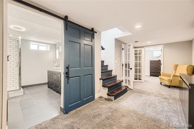 Finished basement with a dark blue barn door, tiled bathroom area, staircase, and yellow armchairs.