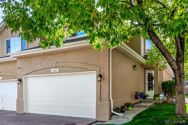 Beige two-story townhouse with a white double garage door beneath a leafy tree.