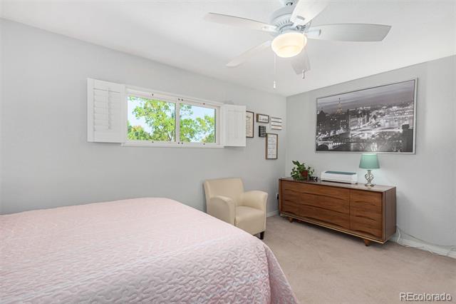 Bedroom with a pink quilted bed, beige armchair, and wooden dresser beneath a cityscape print.
