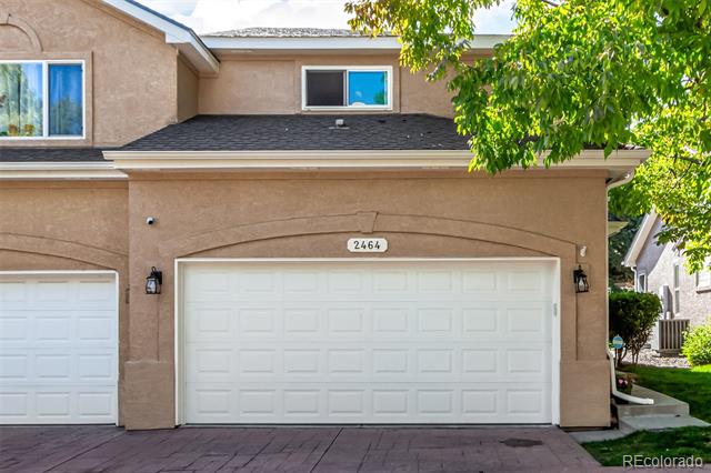 Two-story tan stucco home with a white double garage door and leafy branches overhead.