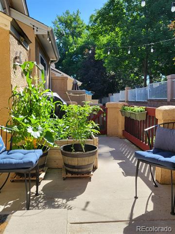 Patio with cushioned chairs, leafy potted plants, and a red railing beside the fenced yard.