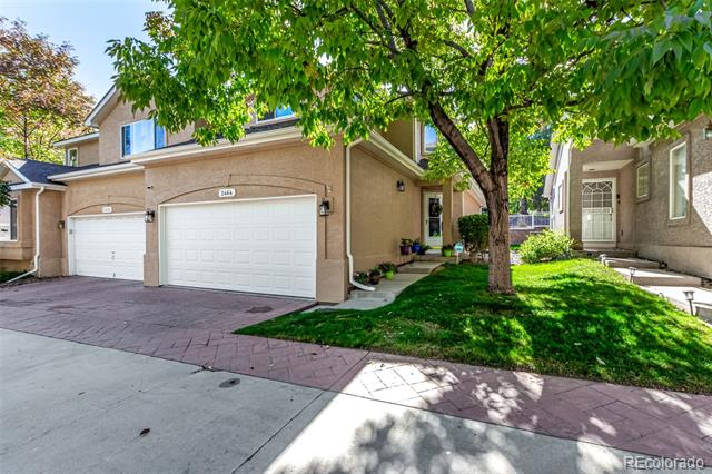Two-story tan townhouse with two white garage doors and a mature tree shading the front lawn.