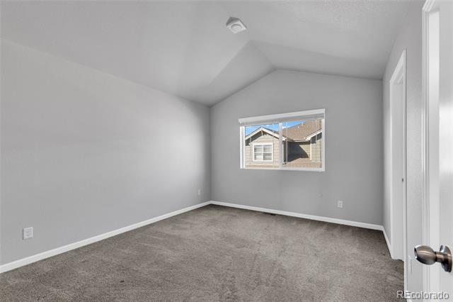 Empty gray bedroom with carpet, a vaulted ceiling, and a window facing a neighboring house.