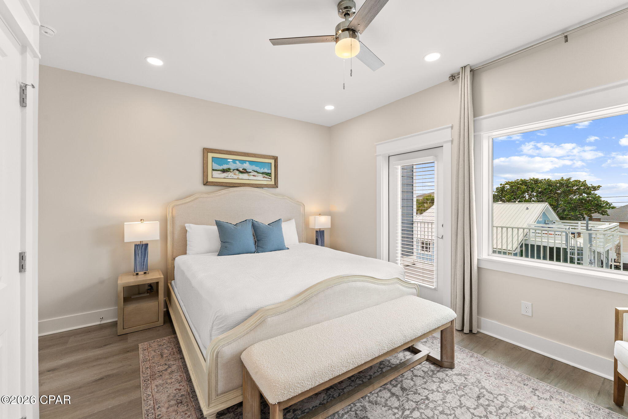 Furnished bedroom with an upholstered bed, blue pillows, and a window overlooking neighboring homes.