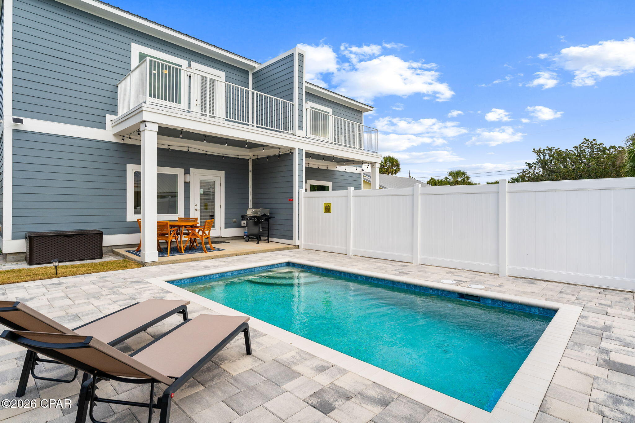 Back patio and pool beside a blue-gray two-story home, with a balcony, white fence, and lounge chairs.