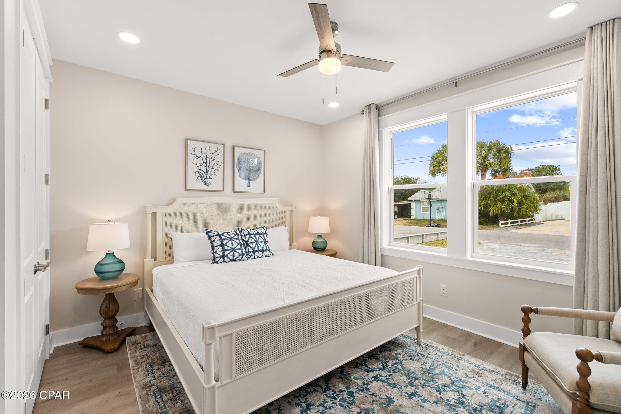 Staged bedroom with a white bed, blue patterned rug, and large windows overlooking palm trees.