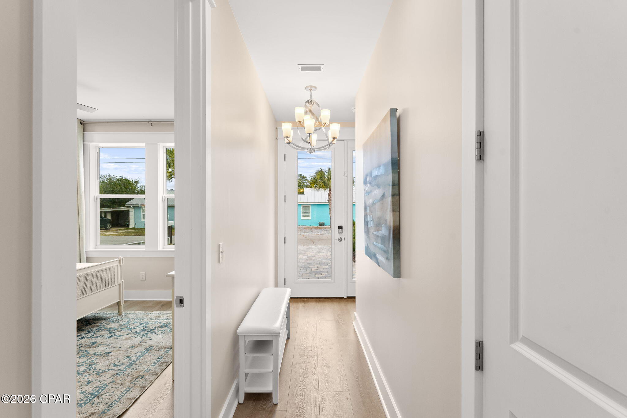 Entry hallway with a white bench, chandelier, and glazed front door facing a turquoise building.