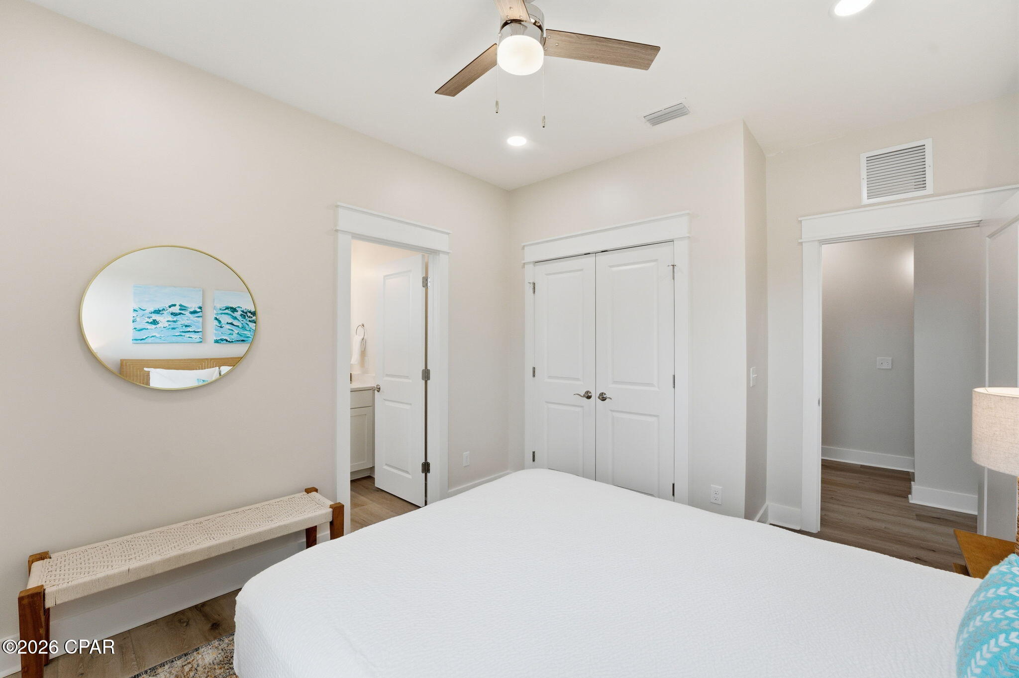 Bedroom with a white bed, ceiling fan, round mirror, and doors to the bathroom and hallway.
