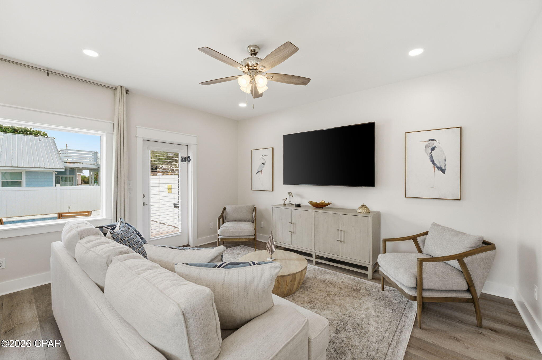 Furnished living room with a white sofa, bird artwork, and a large window overlooking a pool.