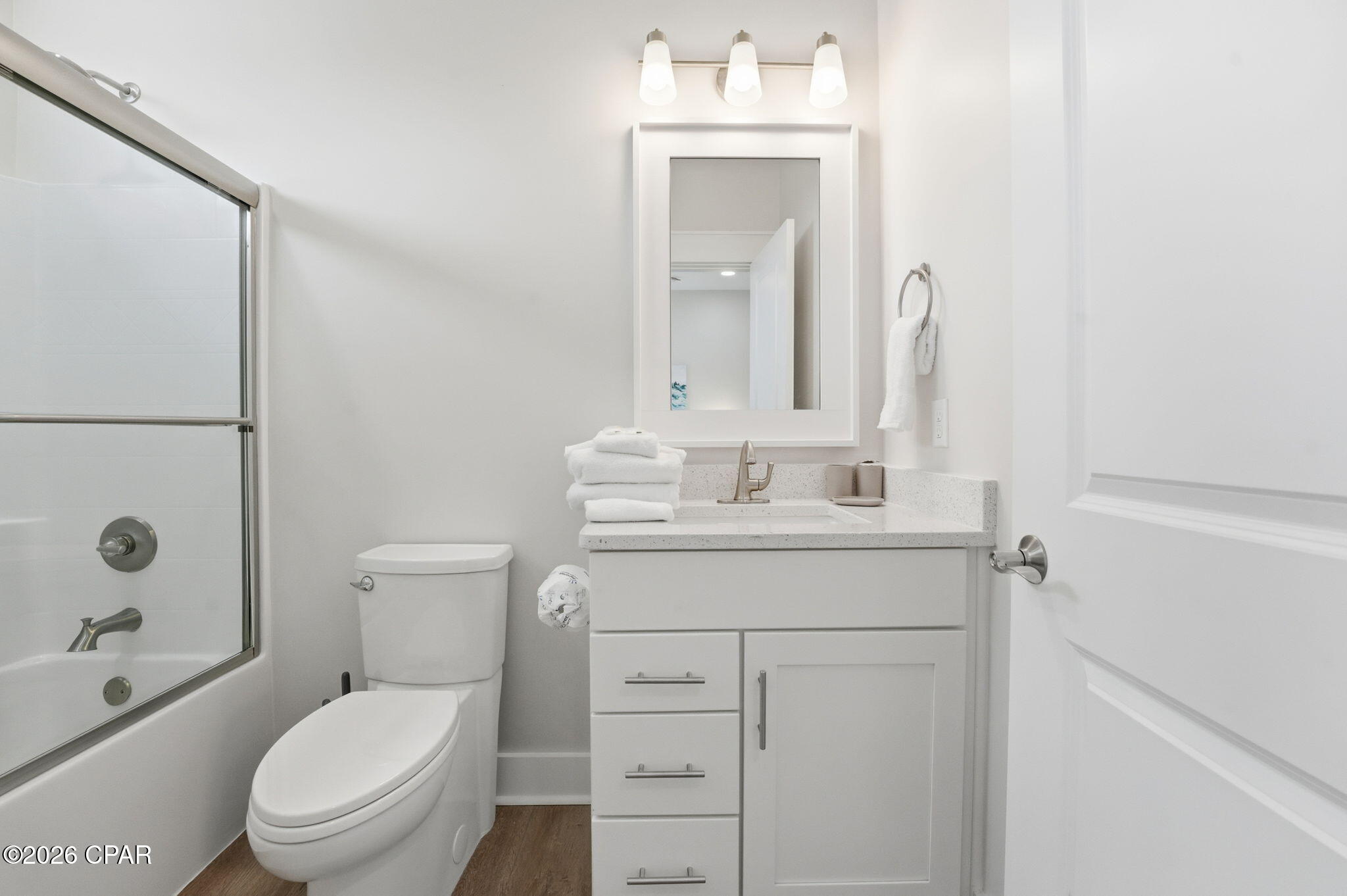 Bright white bathroom with a glass-enclosed tub, toilet, and vanity with folded towels.