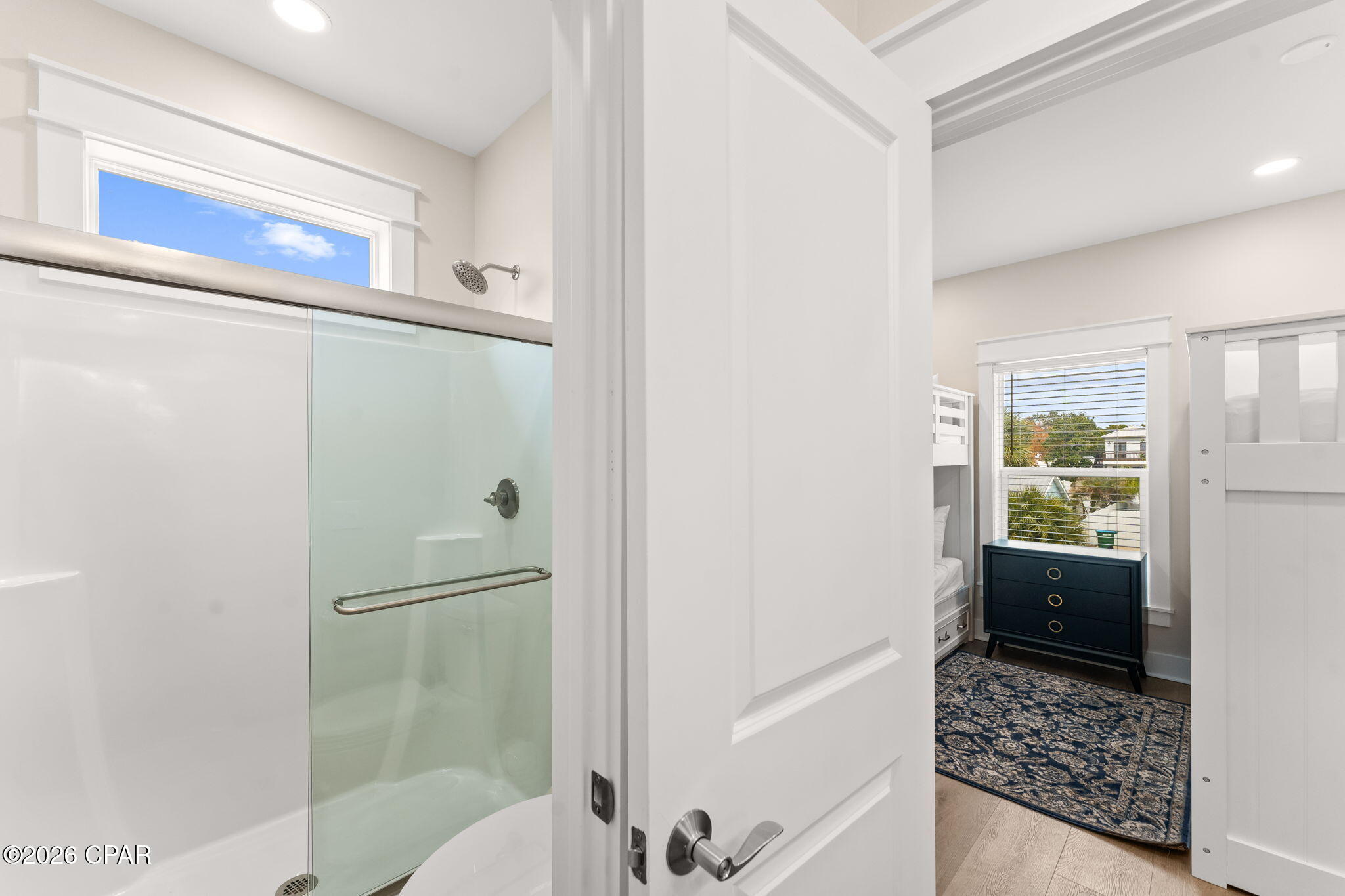 Bathroom with glass shower and high window, opening onto a bedroom with bunk beds and a dark dresser.