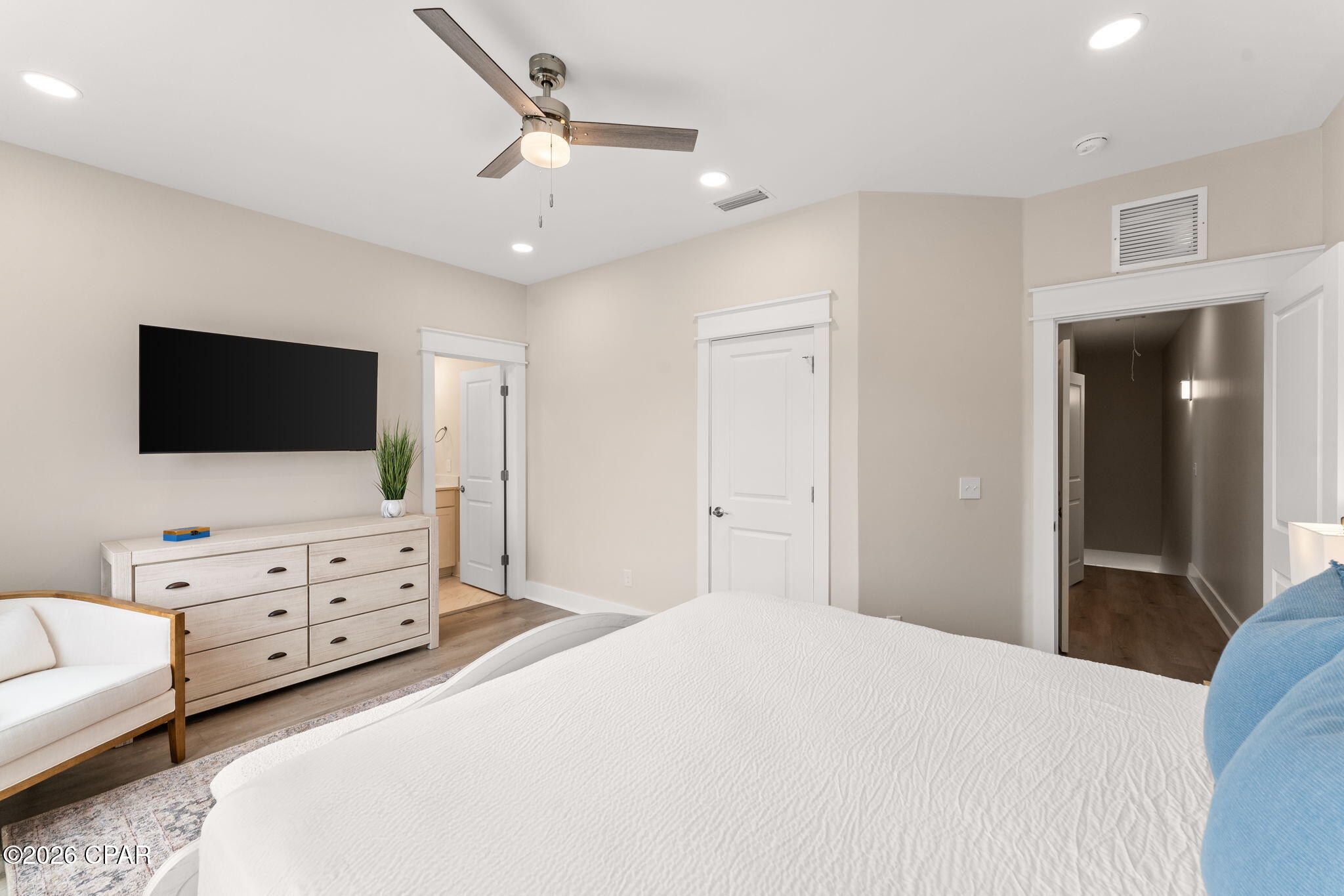 Furnished bedroom with a large white bed, wall-mounted TV, and light wood dresser.