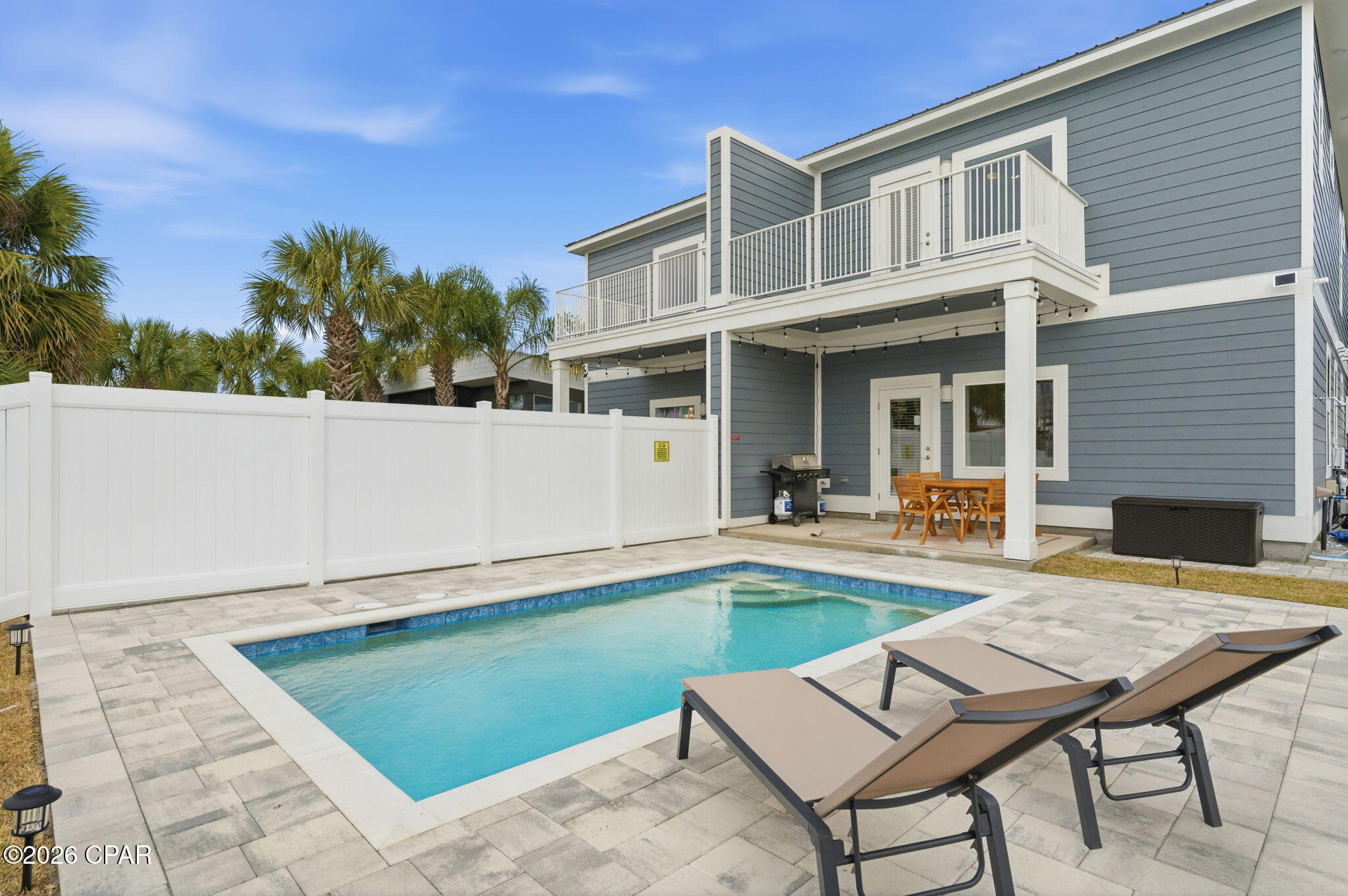 Fenced swimming pool beside a blue-gray two-story home, with patio loungers and a covered dining area.