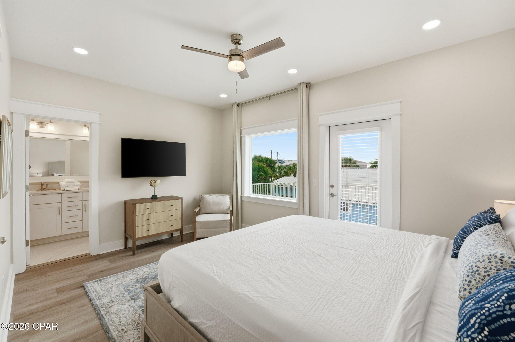 Bedroom with a white bed, wall-mounted TV, ceiling fan, and bathroom doorway.