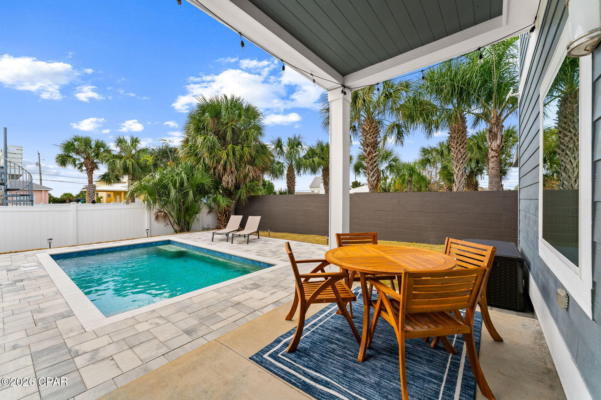 Covered pool patio with wooden dining set, lounge chairs, stone pavers, and palm trees.