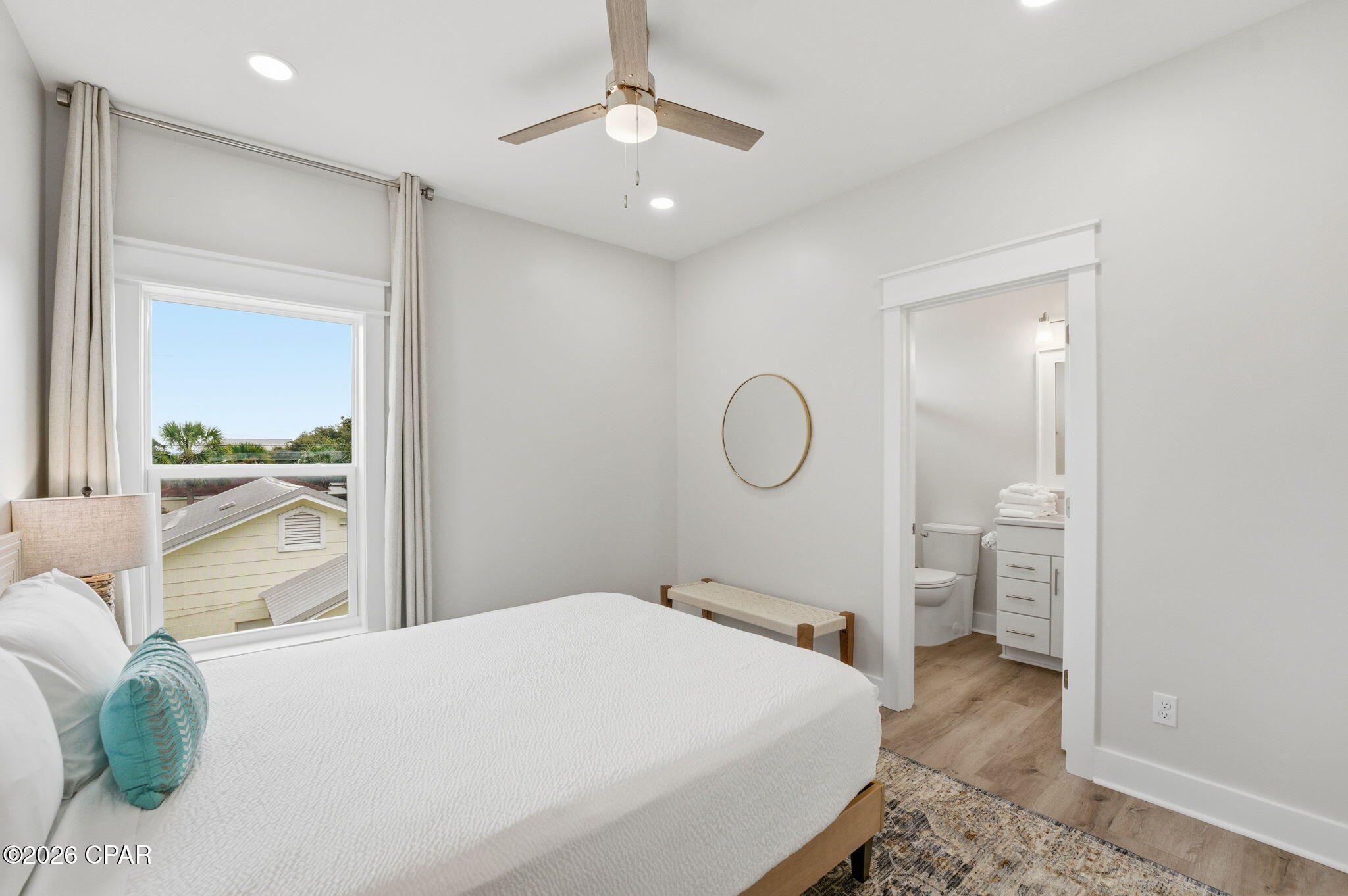 Staged bedroom with a white bed, turquoise pillow, and ensuite bathroom visible through the open doorway.
