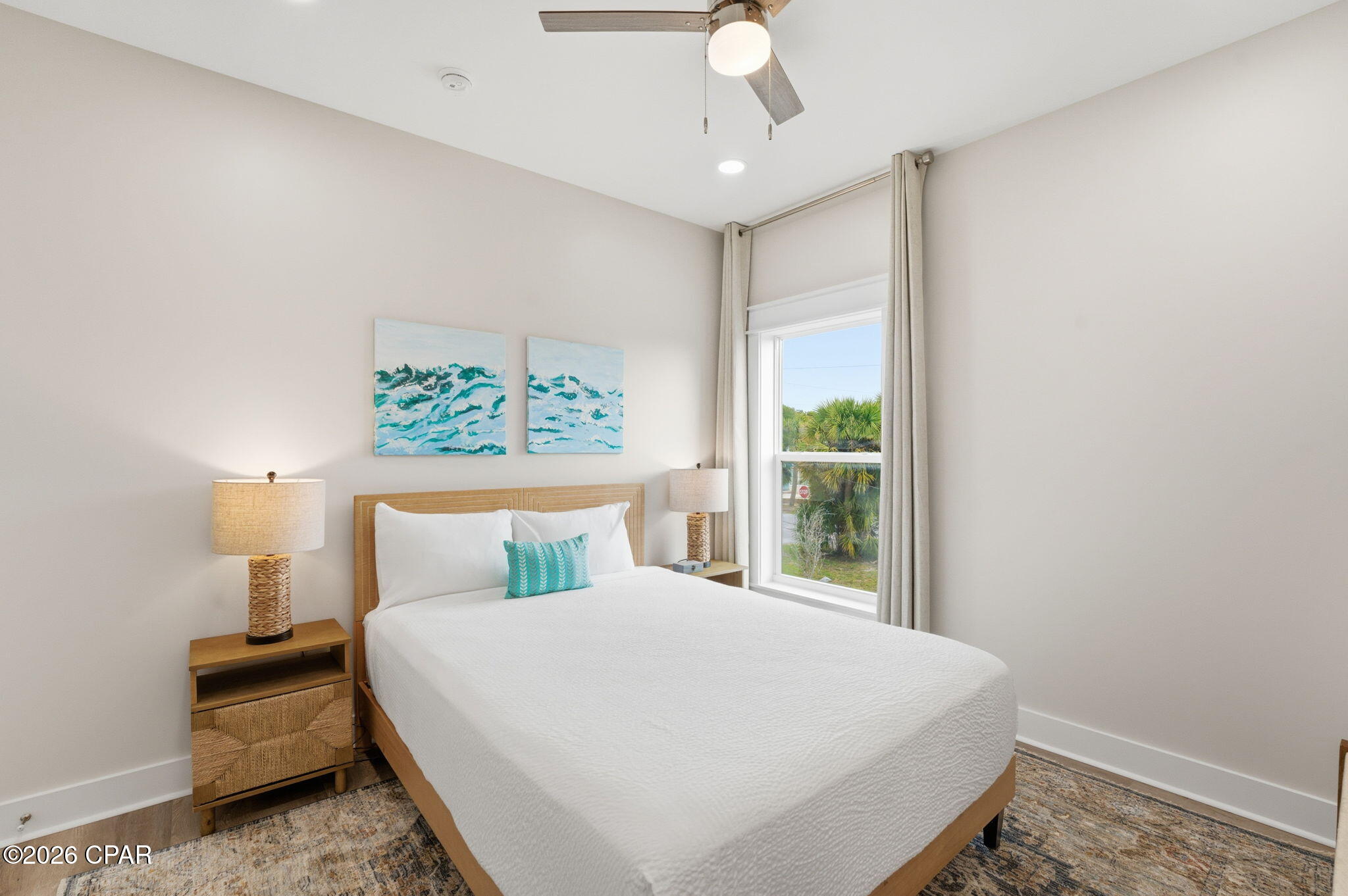 Furnished bedroom with a white bed, turquoise ocean artwork, and a window overlooking palm trees.