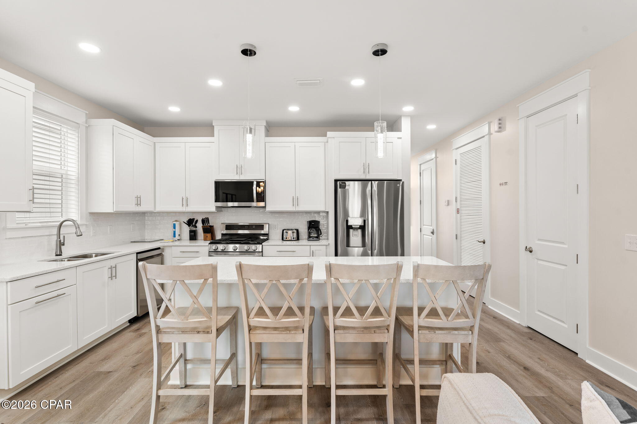 Bright white kitchen with a large island, four wooden bar chairs, and stainless steel appliances.