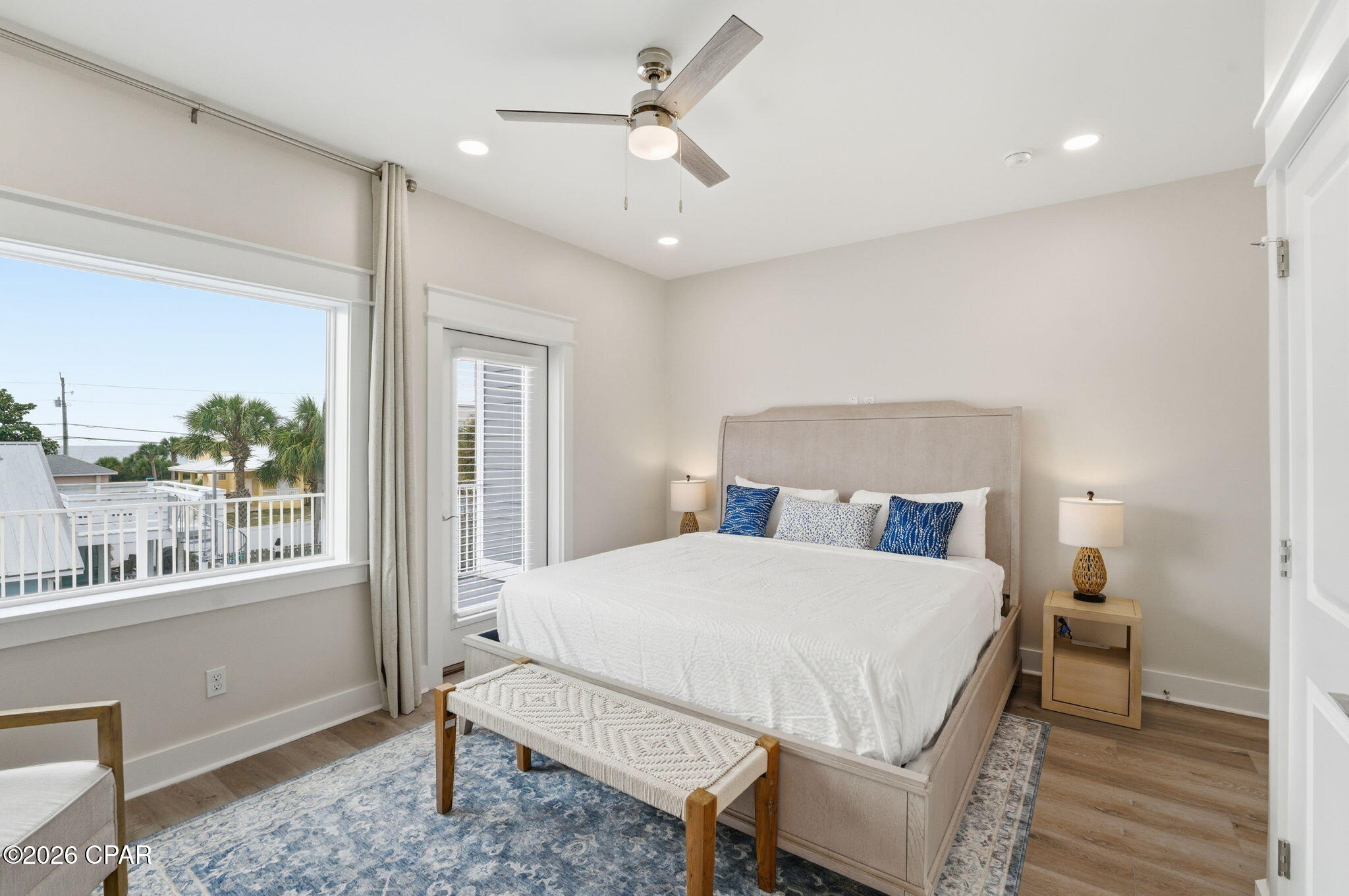 Bedroom with a king bed, blue accents, and a large window overlooking palm trees and nearby homes.