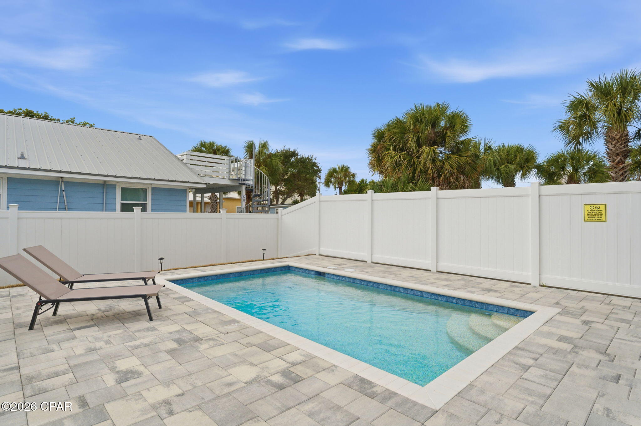Outdoor swimming pool with paver patio, two chaise lounges, and a tall white privacy fence.