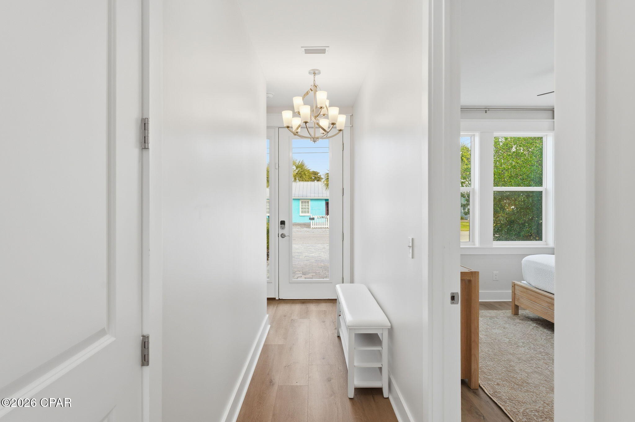 Entry hallway with a chandelier, white bench, and glass-paneled front door beside a bedroom.