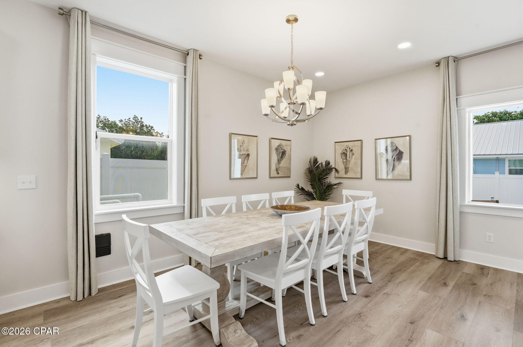 Dining room with a pale wood table, white chairs, chandelier, and large curtained windows.