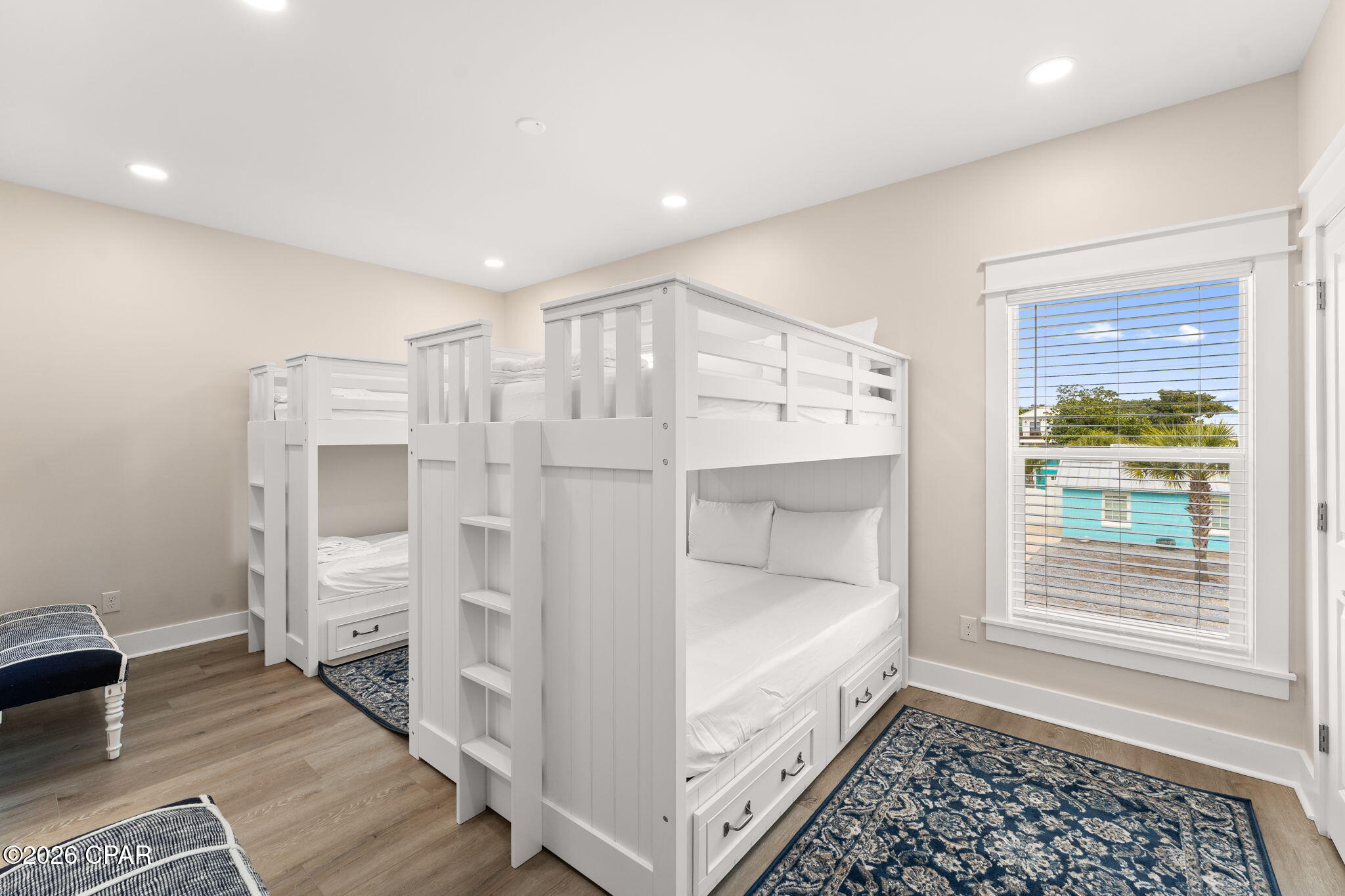 Bedroom with two white bunk beds, built-in drawers, blue rugs, and a window overlooking a palm tree.