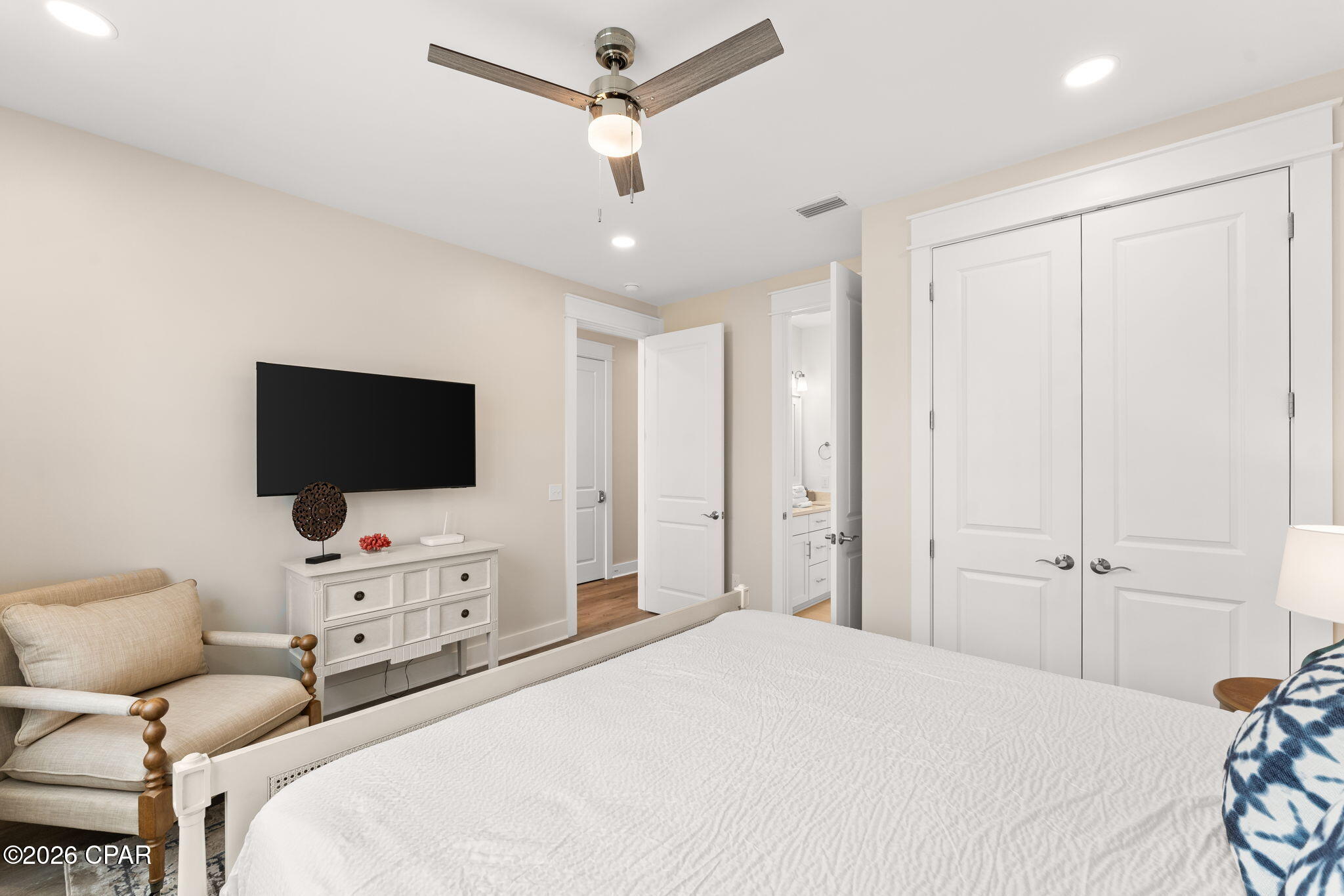Furnished bedroom with a white bed, beige armchair, and wall-mounted television beside open bathroom and closet doors.