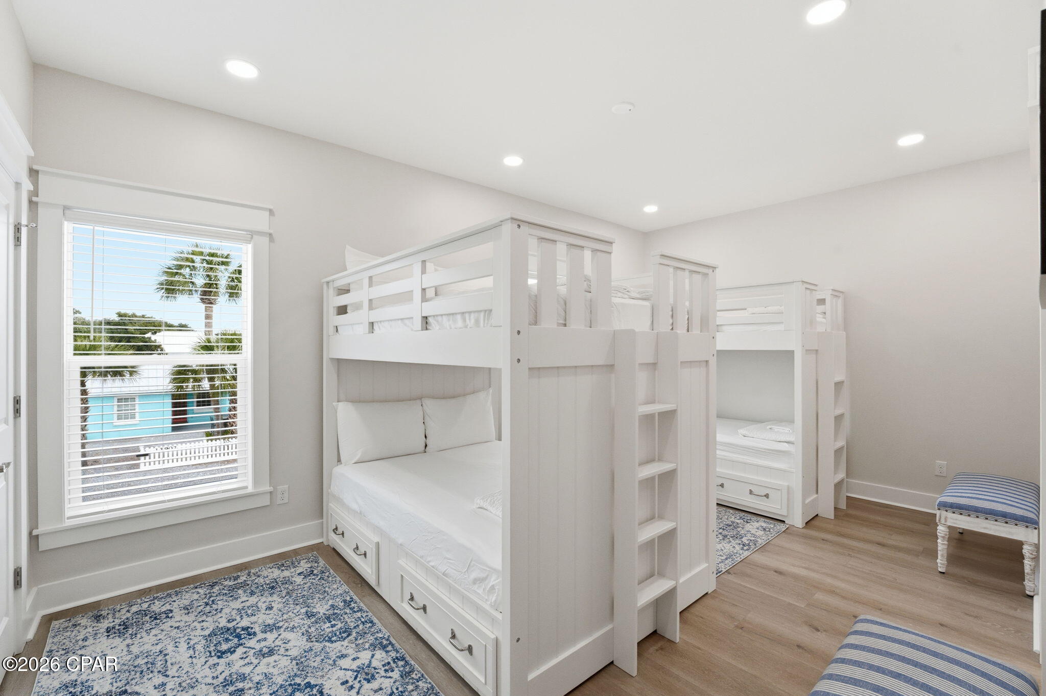Bedroom with white built-in bunk beds, storage drawers, blue rugs, and a window overlooking palm trees.