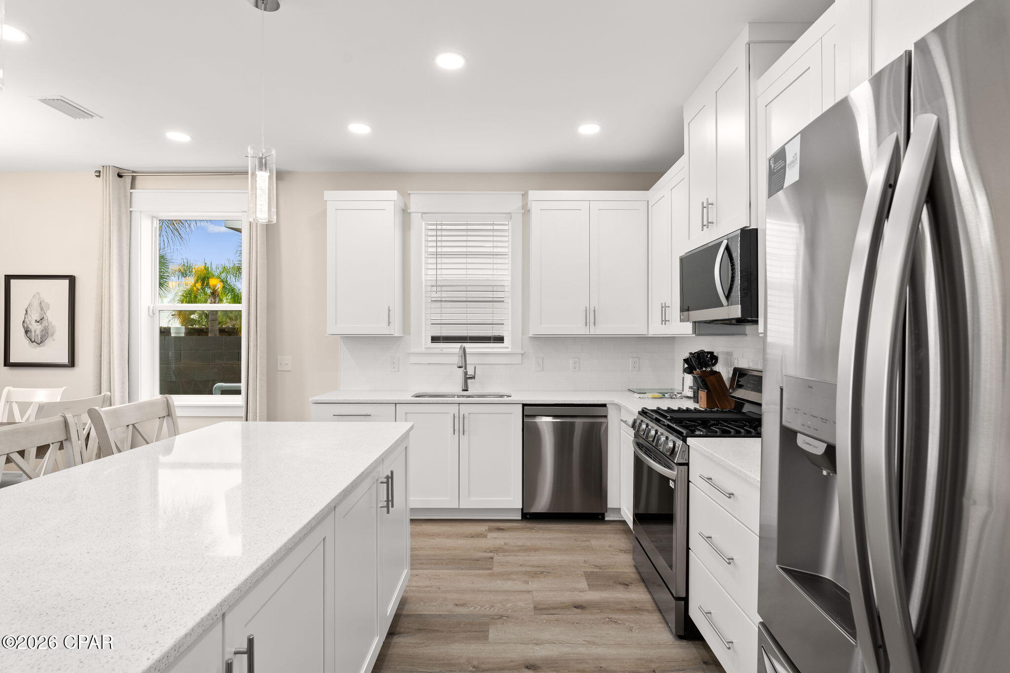 Bright white kitchen with a large quartz island, stainless steel appliances, and a window overlooking palm trees.