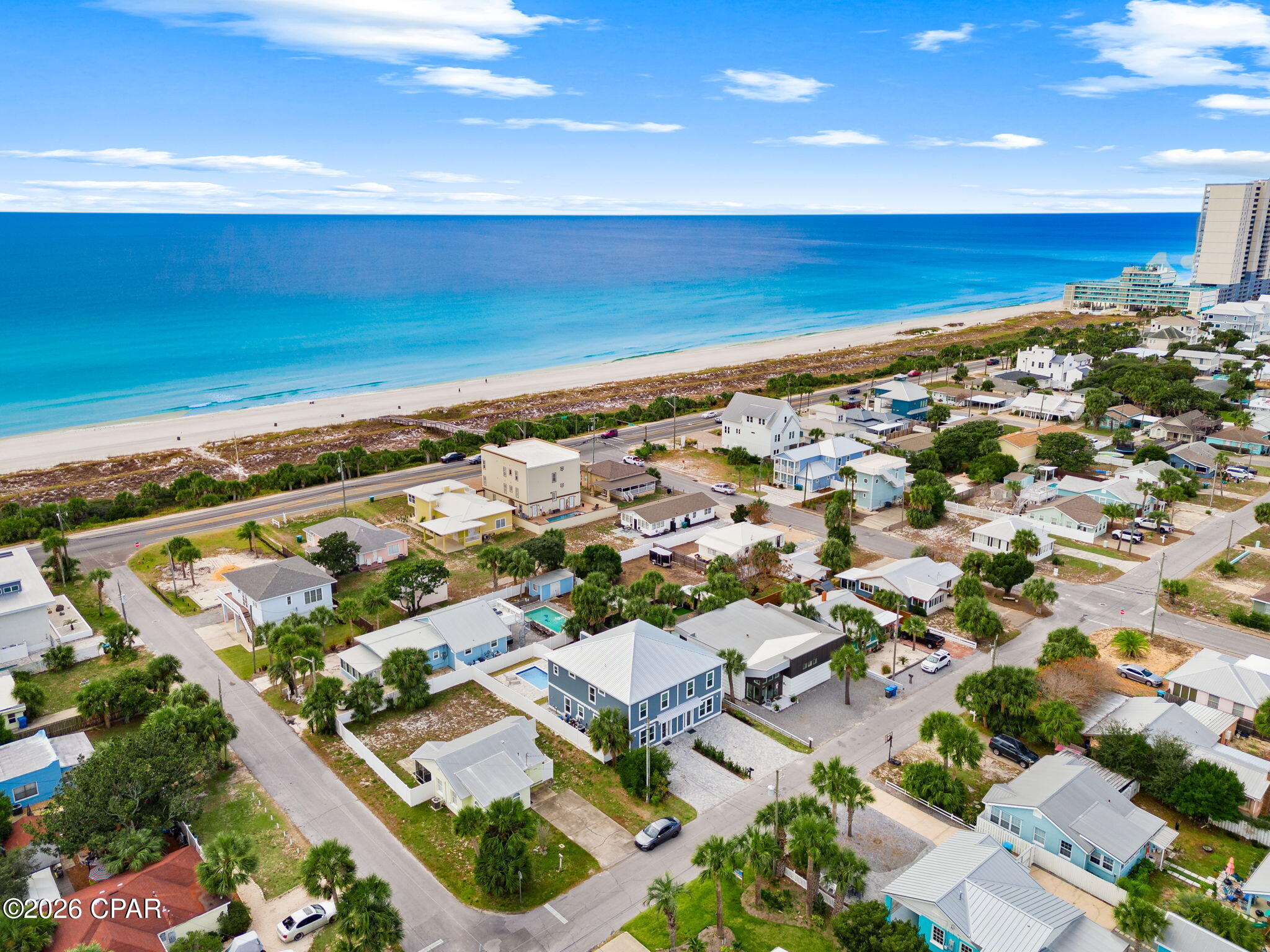 Aerial view of coastal homes beside a sandy beach and turquoise ocean, with a blue house near the foreground.