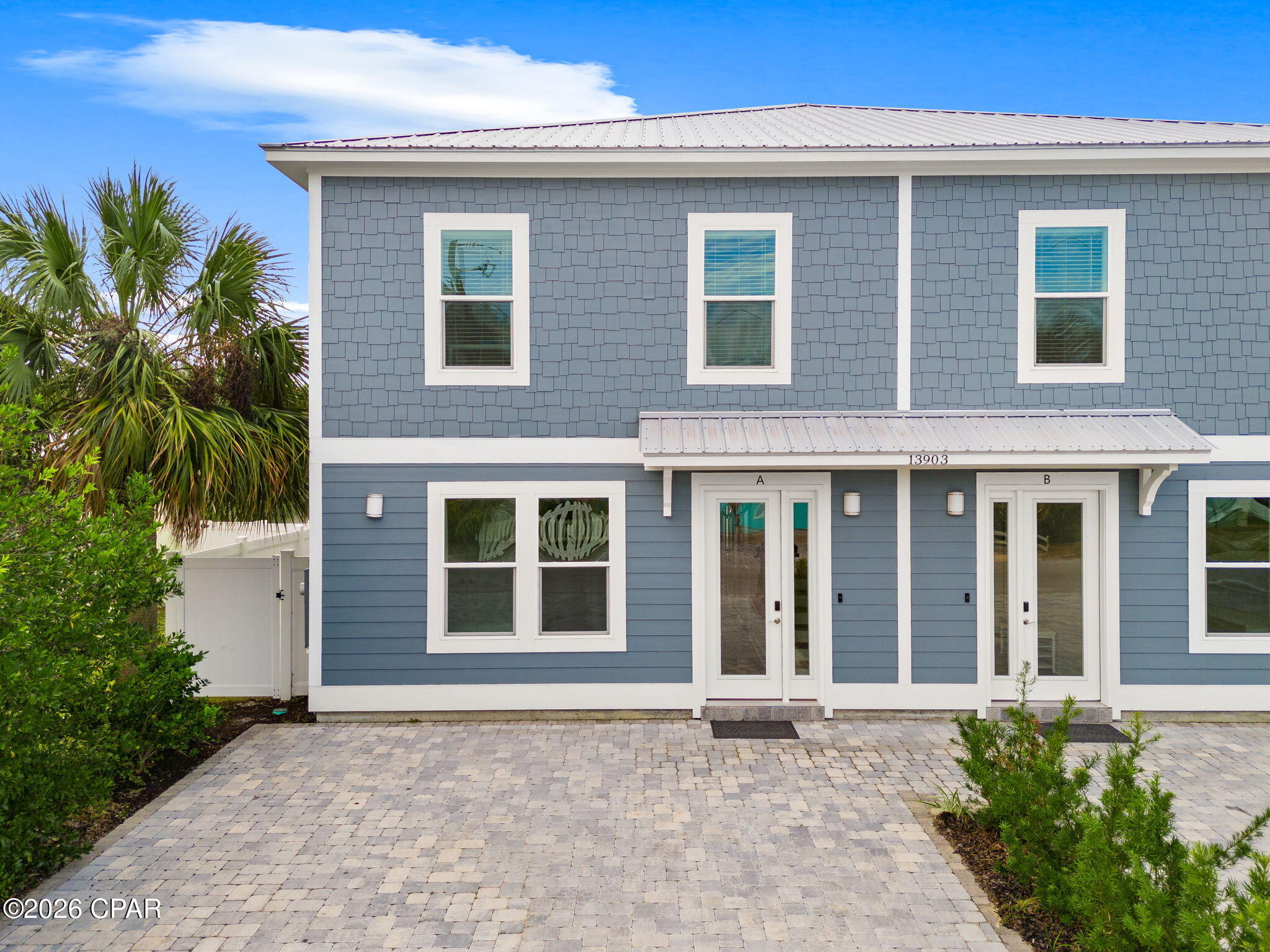 Blue two-story duplex with separate A and B entrances, a broad paver driveway, and palm trees.