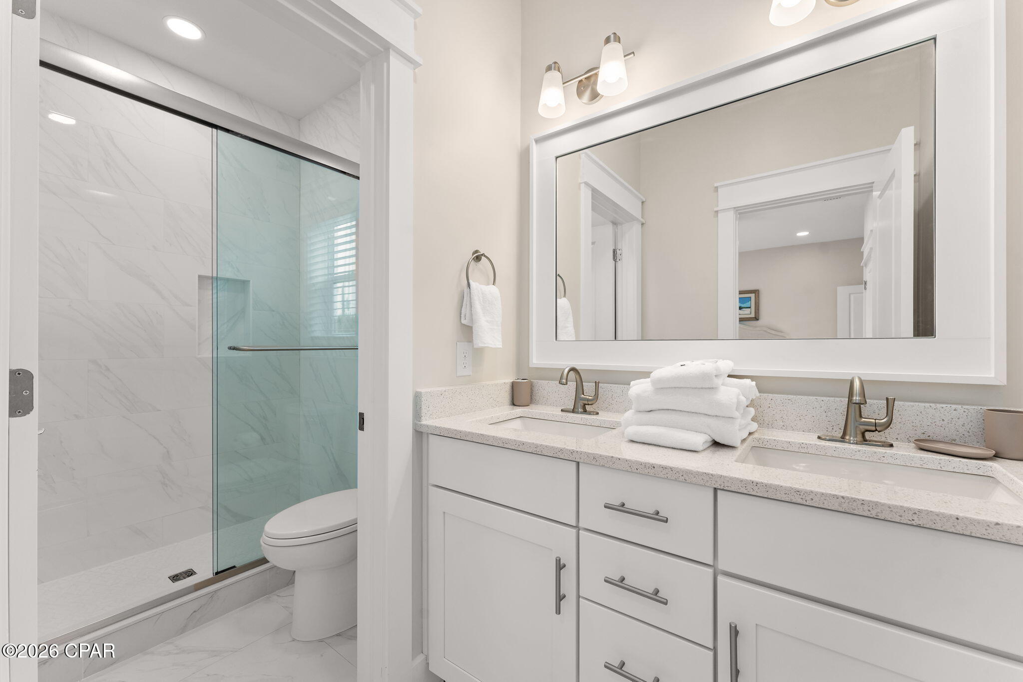 Bathroom with a marble-look glass shower and white double vanity with folded towels.