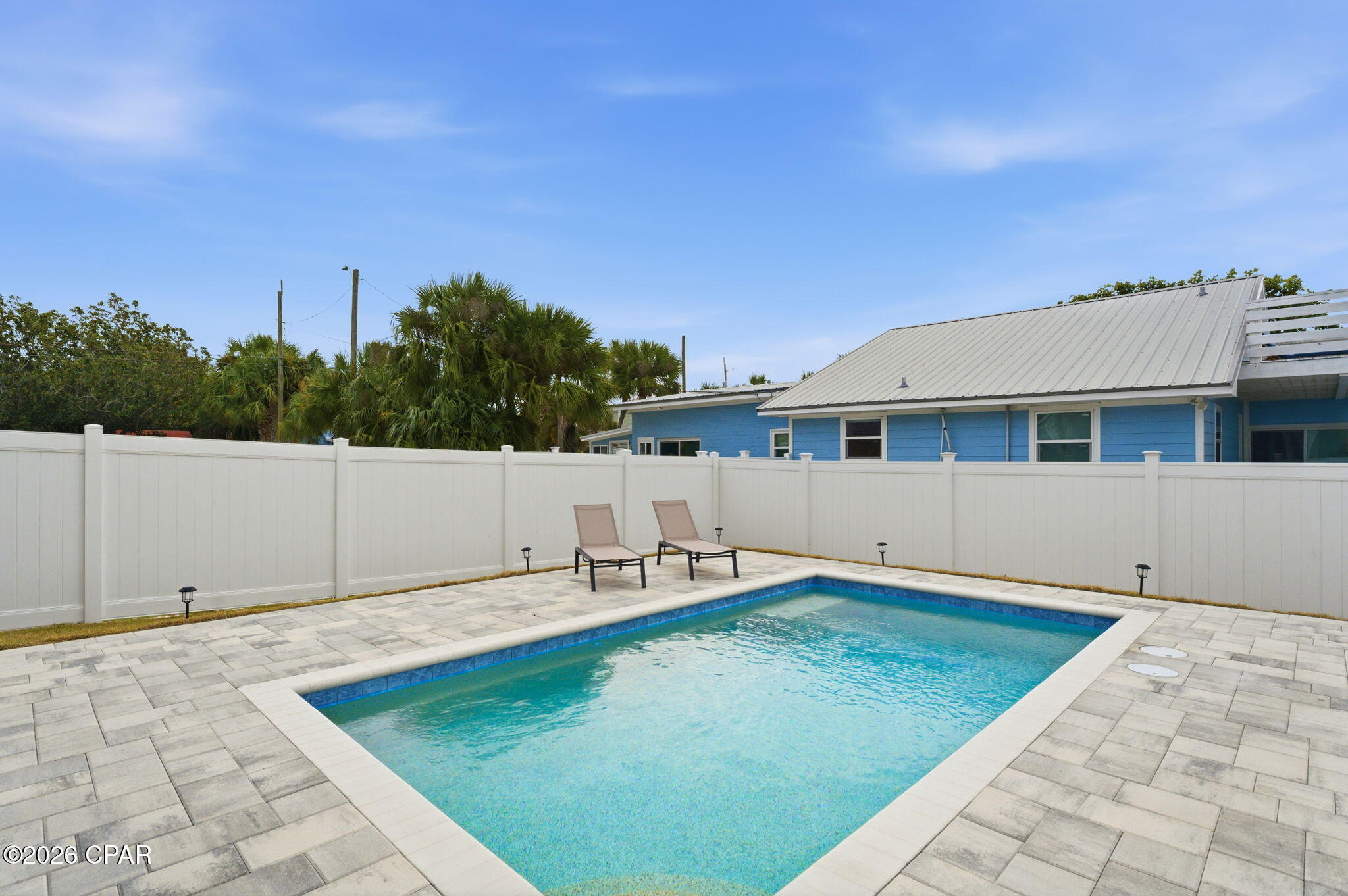 Outdoor pool with a paved deck, white privacy fence, and two lounge chairs beside a blue house.