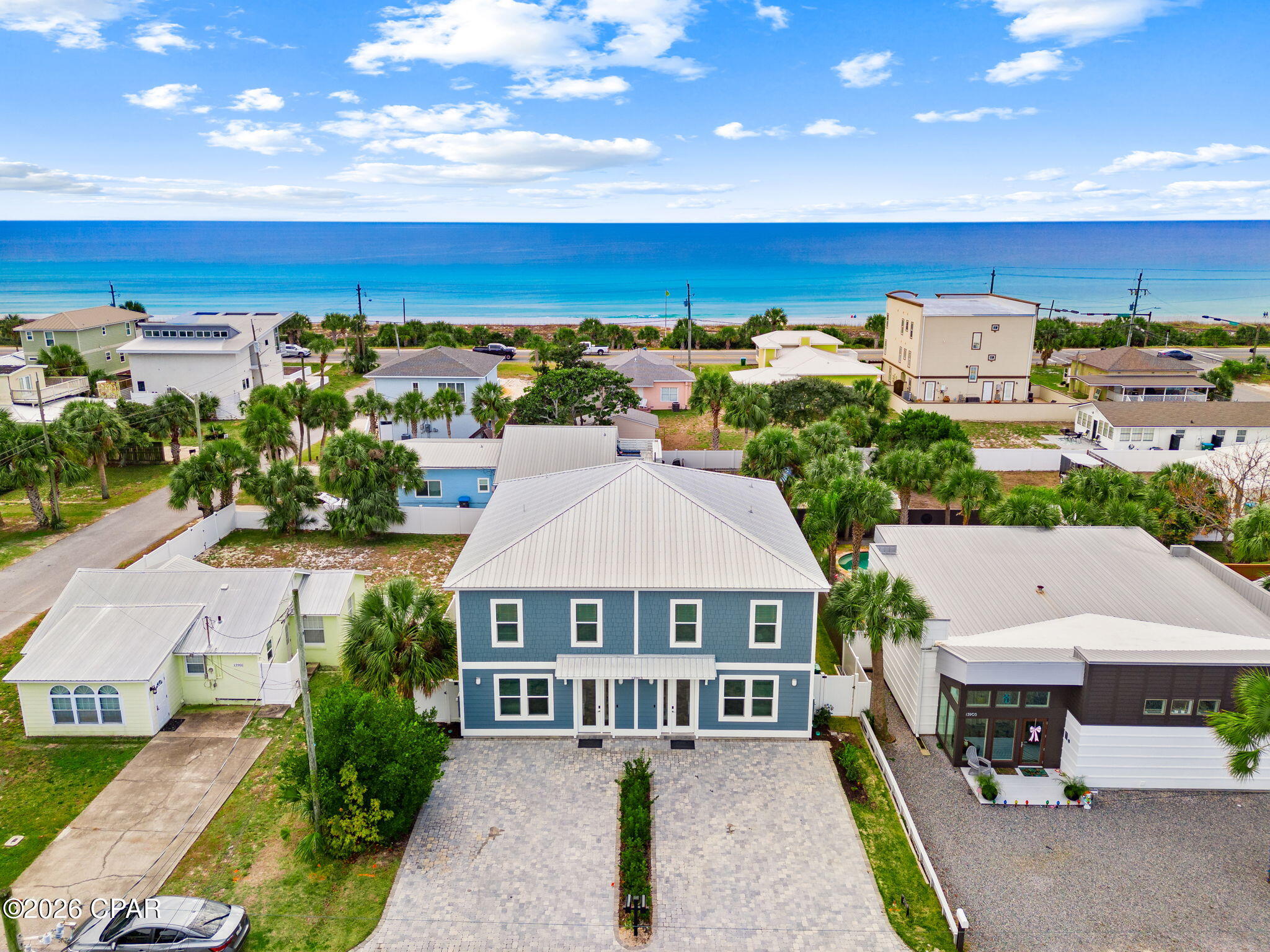 Blue two-story coastal home with a paved driveway, palm trees, and ocean behind it.