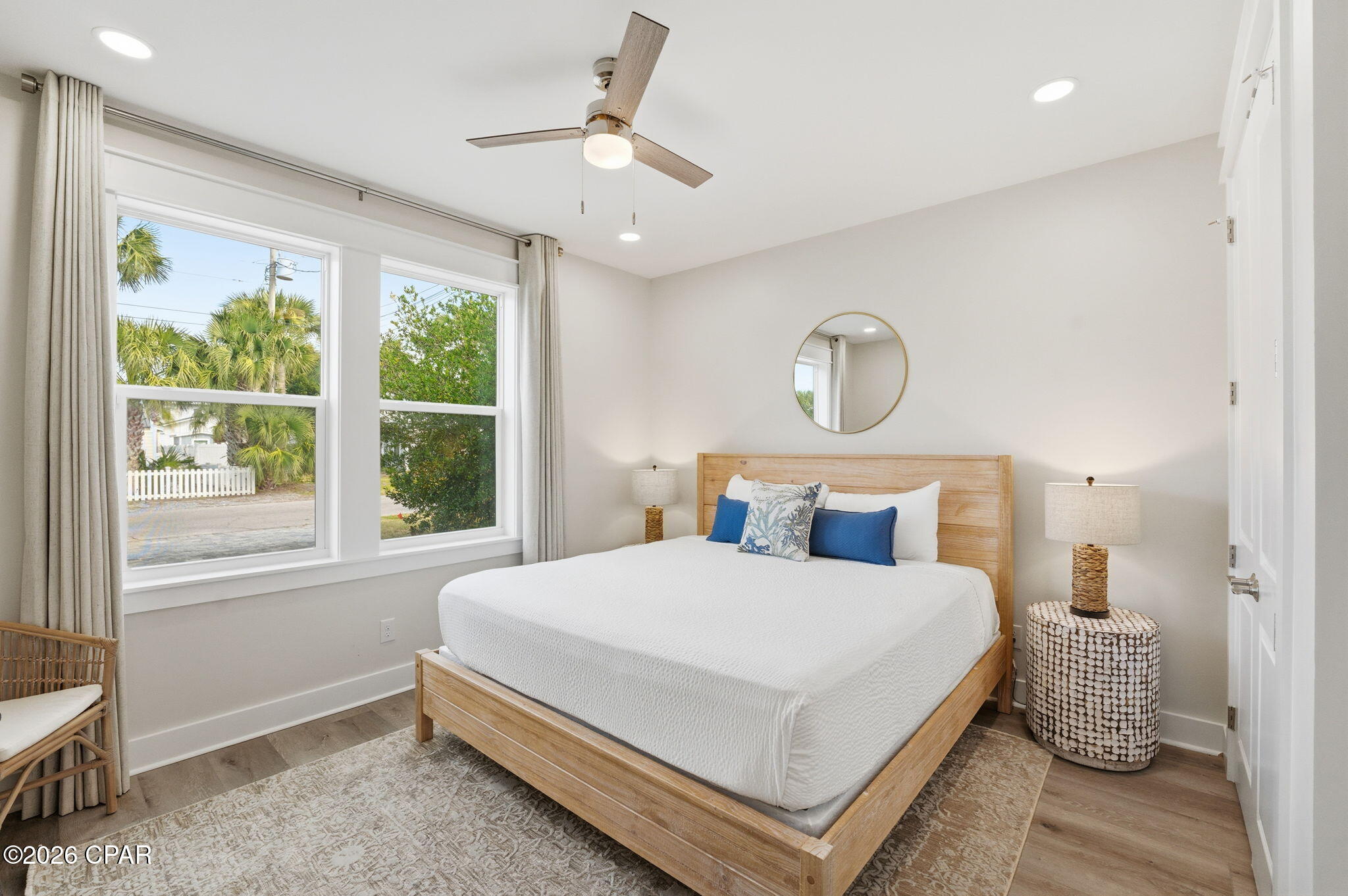 Bedroom with a light wood bed, blue accent pillows, and windows overlooking palm trees.