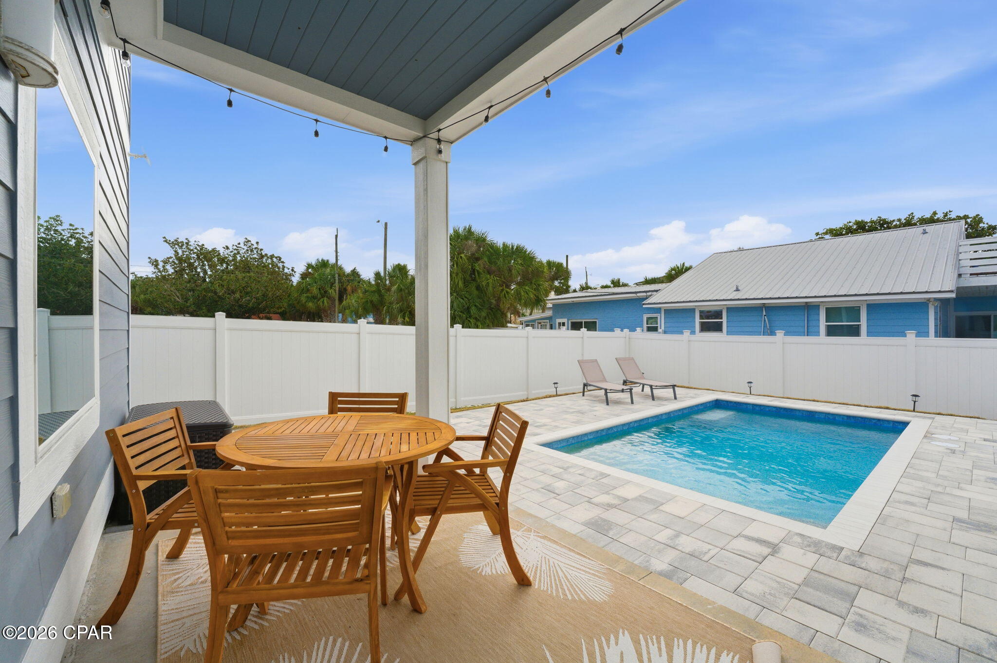 Backyard pool beside a covered patio with wooden dining furniture and two loungers.