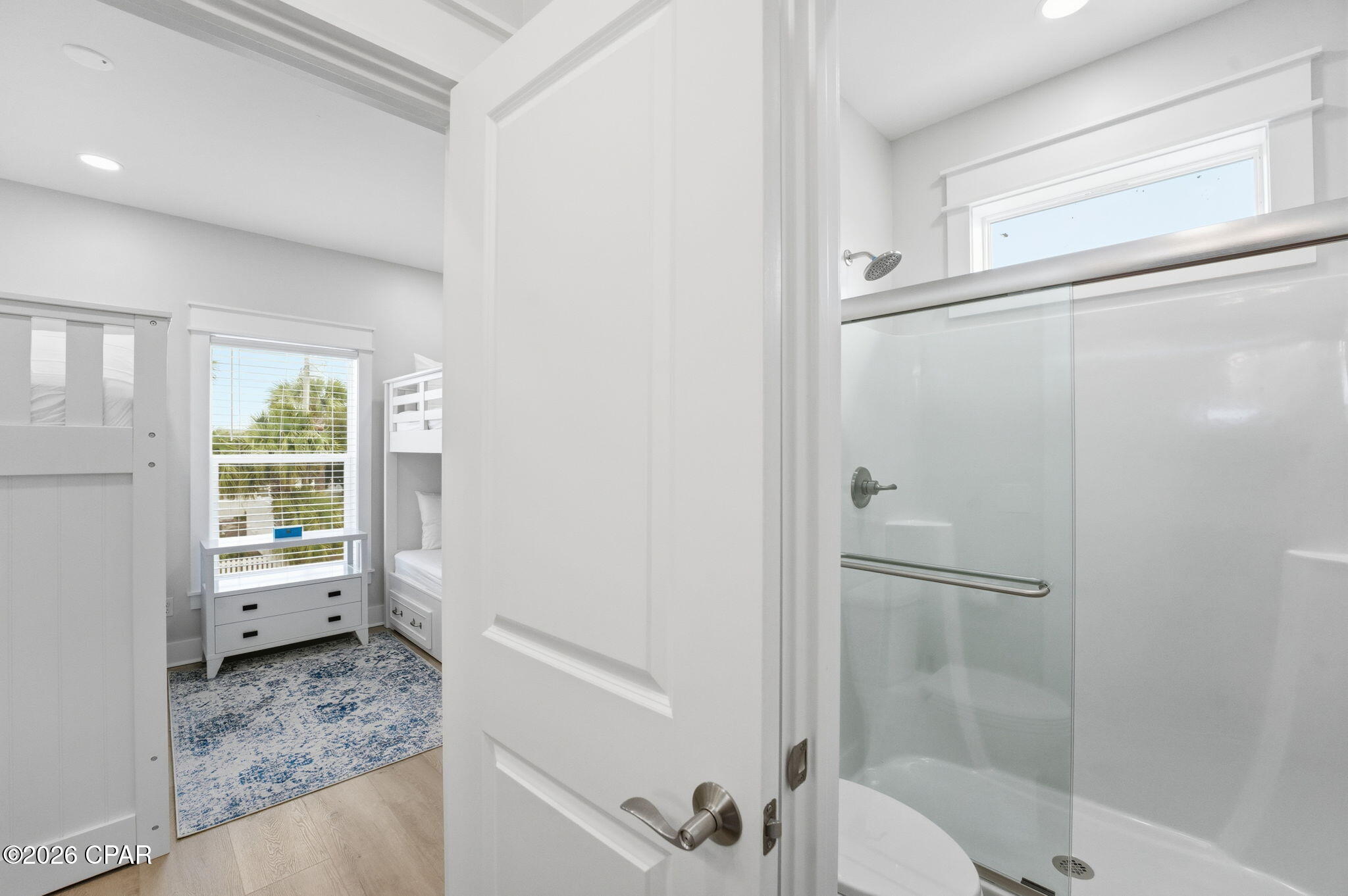 Bathroom with a glass shower, viewed beside a bunk room with a blue patterned rug.