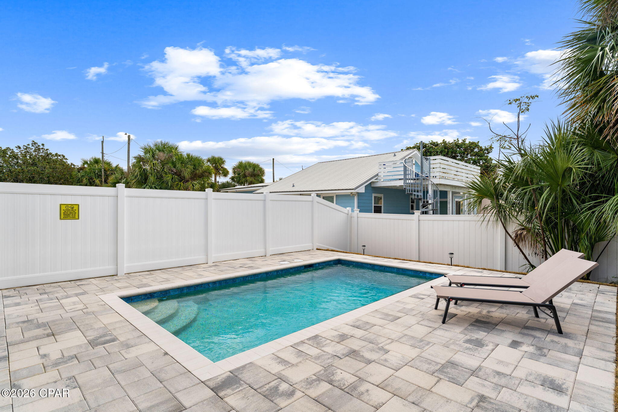Outdoor pool with two lounge chairs, pale paver decking, a white privacy fence, and tropical palms.