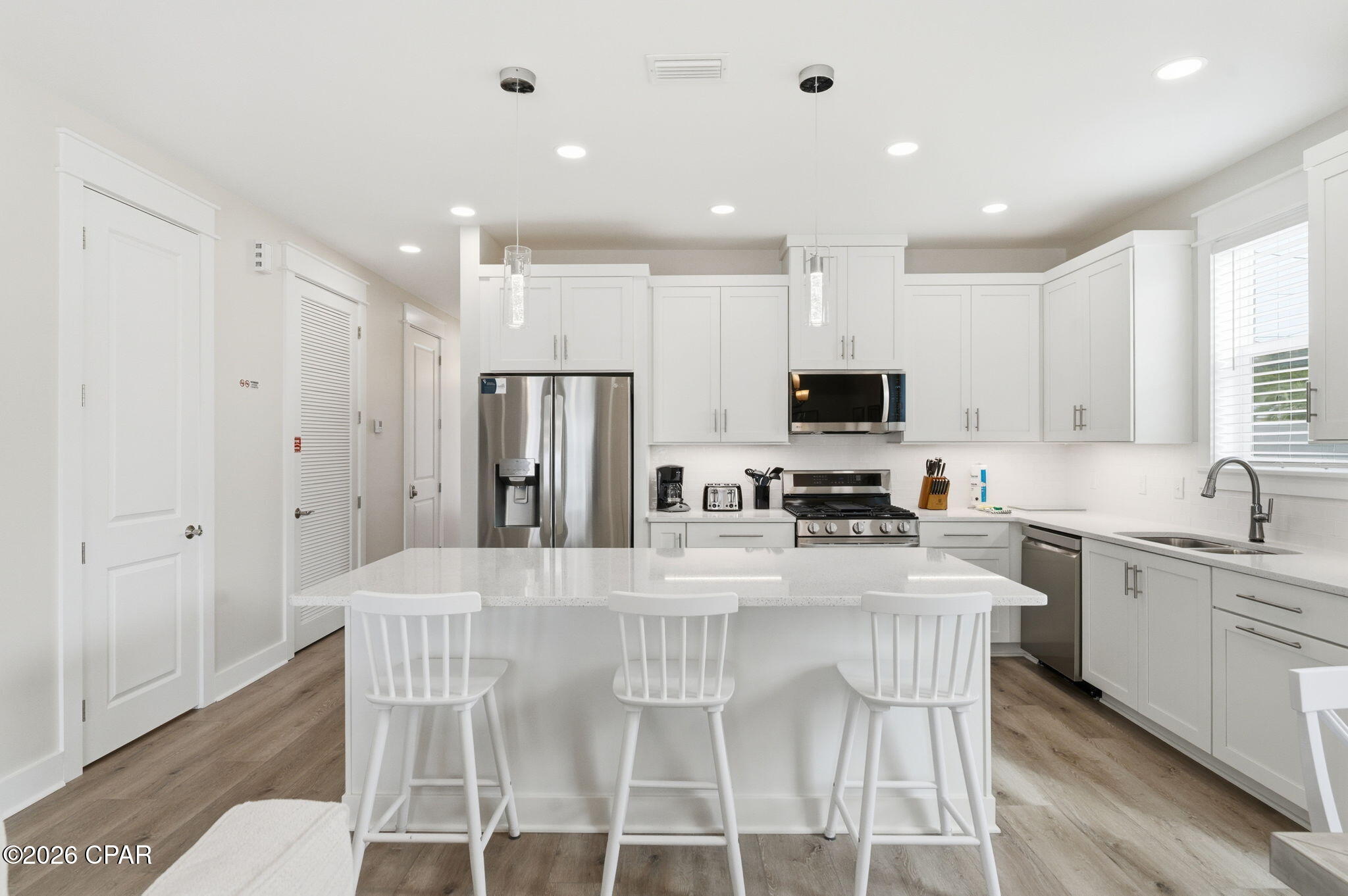 Bright white kitchen with a large island, three stools, and stainless steel appliances.