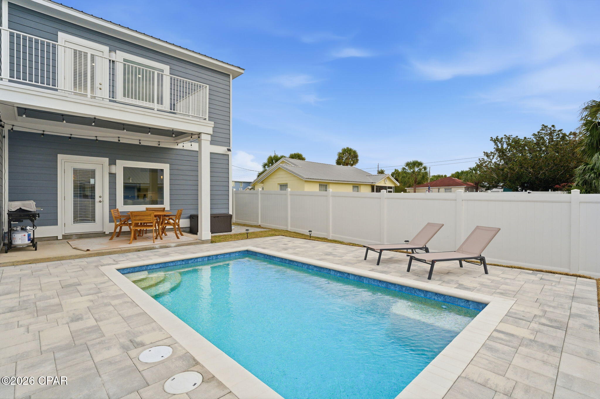 Backyard swimming pool beside a gray two-story house, with a fenced patio and lounge chairs.