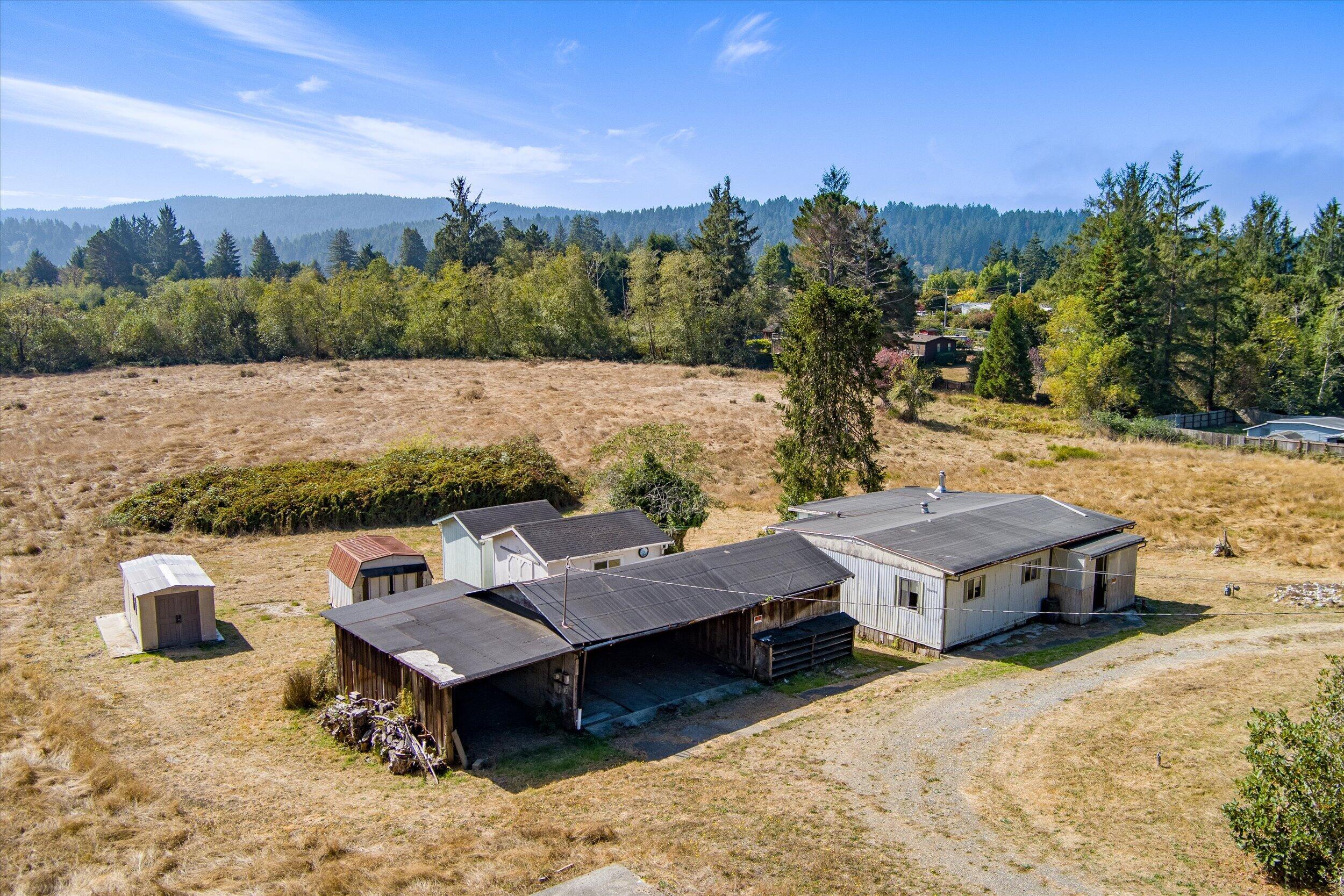 Aerial view of rural outbuildings in a dry field bordered by trees and wooded hills.