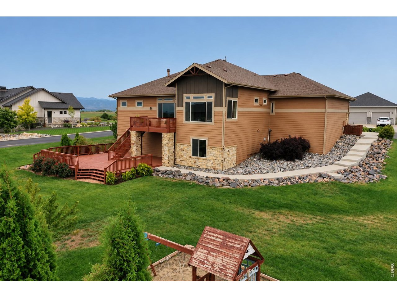 Rear of a tan home with stone accents, a raised redwood deck, and a broad lawn with a playset.