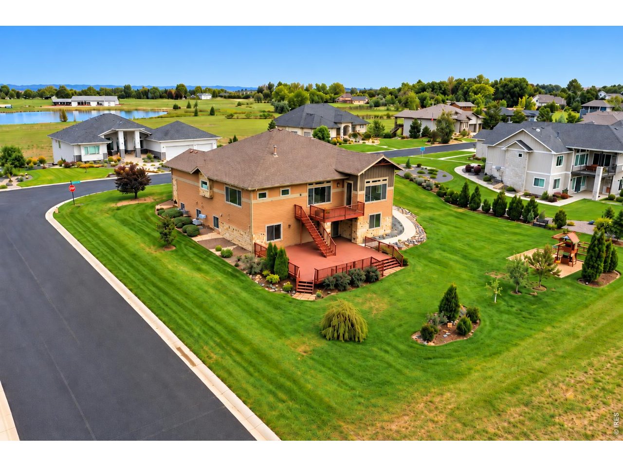Aerial view of a two-story home with a broad deck and landscaped lawn beside a curving neighborhood road.