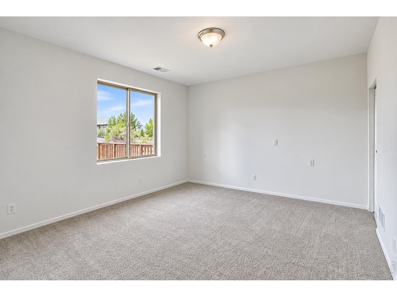 Empty carpeted room with a large window overlooking a wooden deck railing and greenery.