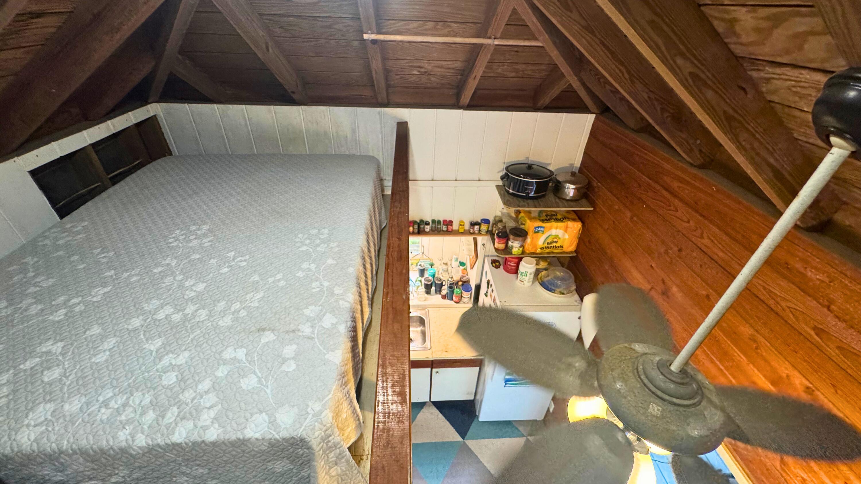 Furnished loft bedroom with exposed rafters, a low bed, kitchenette, and ceiling fan.