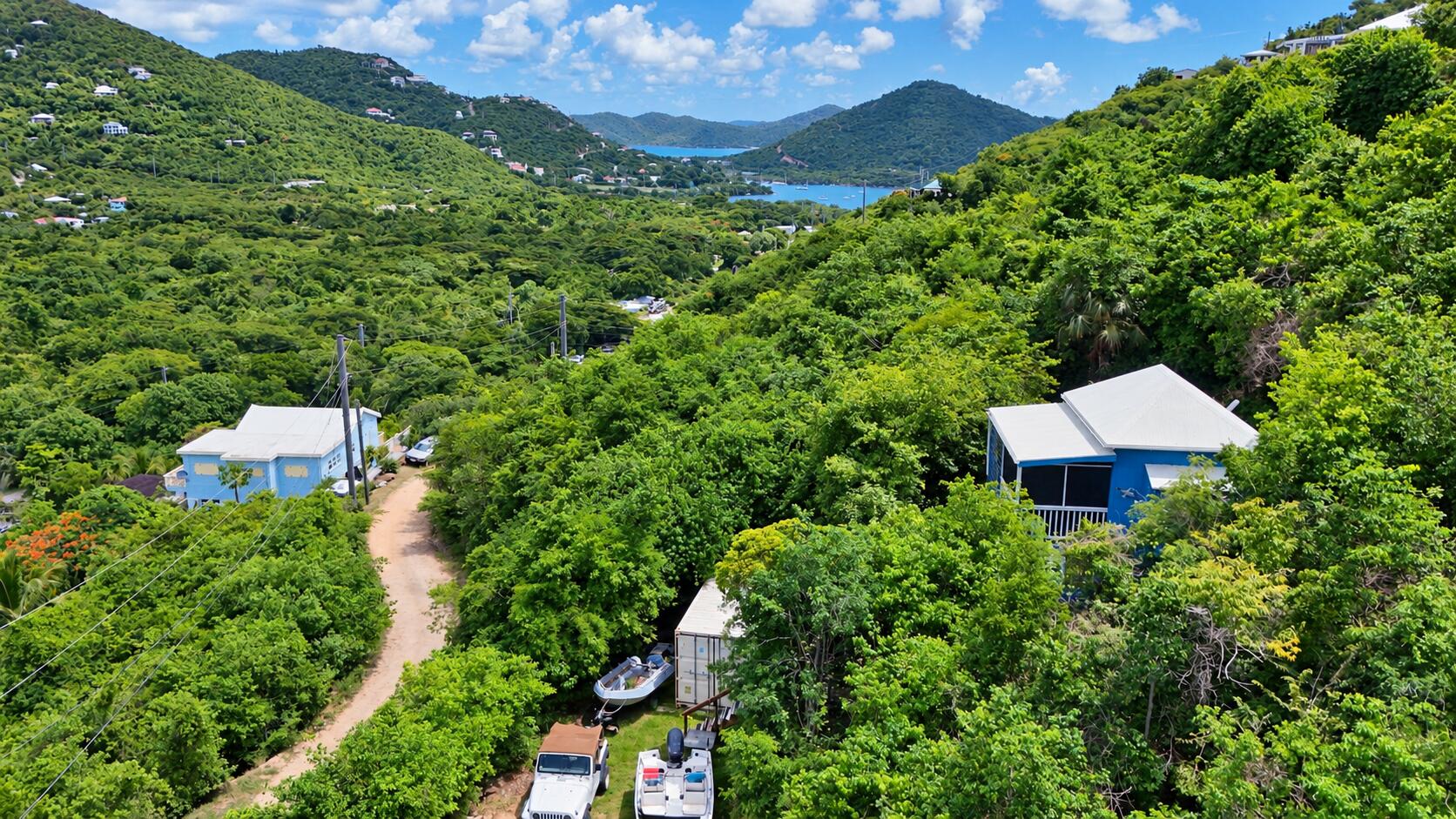 Aerial hillside view with blue houses, a winding dirt road, boats, and a bay beyond the green hills.